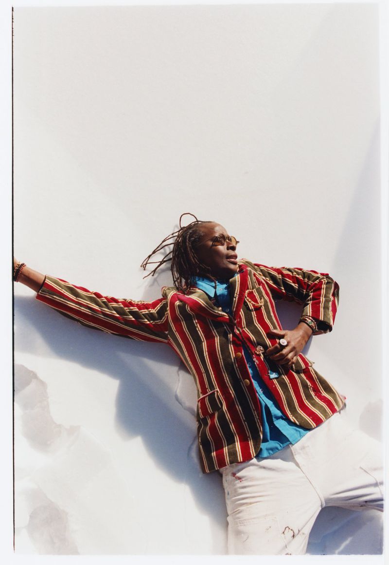 A Black person with dreadlocks wearing a red-and-green striped blazer and white trousers reclines against a sunlit white surface.