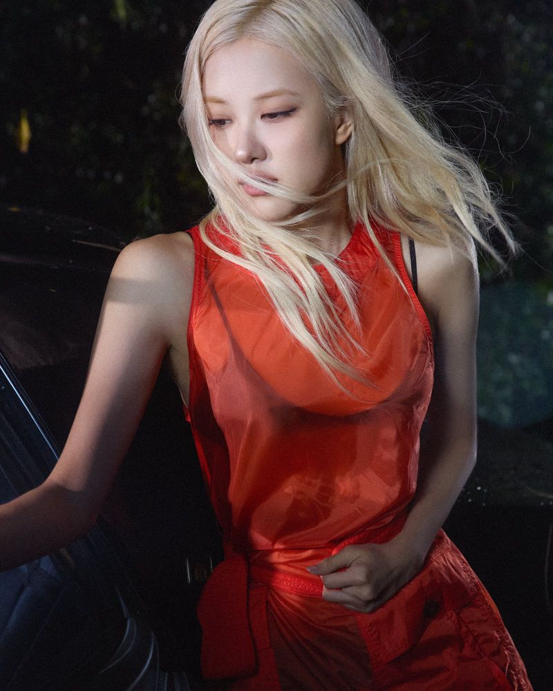 Rosé with windblown blonde hair wearing a bright red sheer sleeveless top against a dark outdoor backdrop at night.