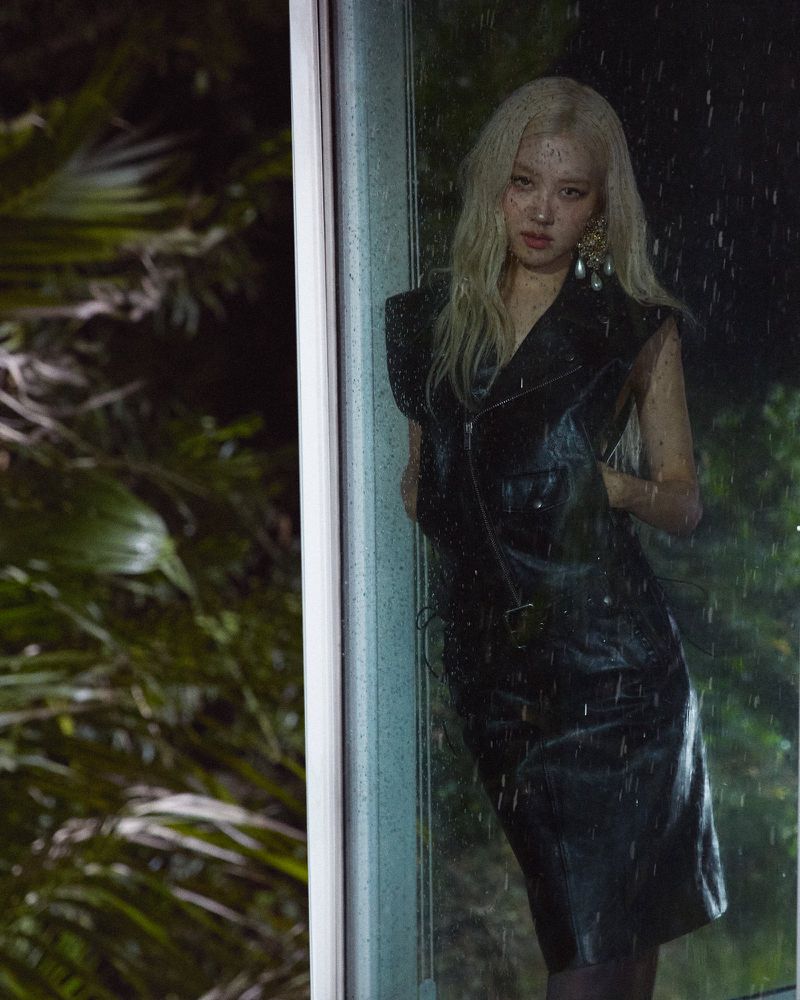 Adult female with blonde hair poses in a sleeveless black dress while standing near a glass door at night in a nighttime outdoor setting.