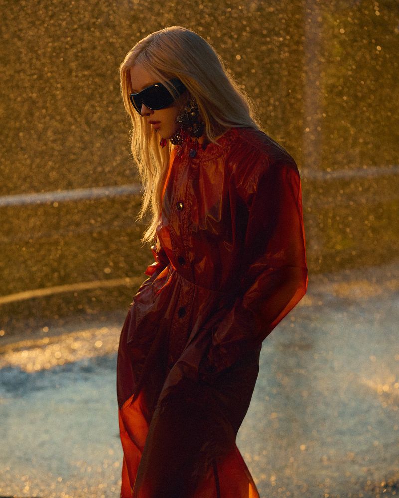 Rosé wearing a translucent red trench coat and black shield sunglasses against a backlit golden outdoor background.