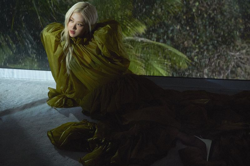 Rosé in a voluminous olive green ruffled gown reclining indoors before a rain-streaked window with palm trees in the background.