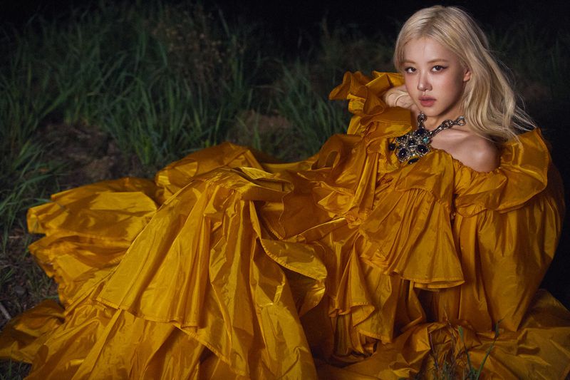 Rosé Park reclining in an voluminous golden yellow ruffled off-shoulder gown in a dark field at night.