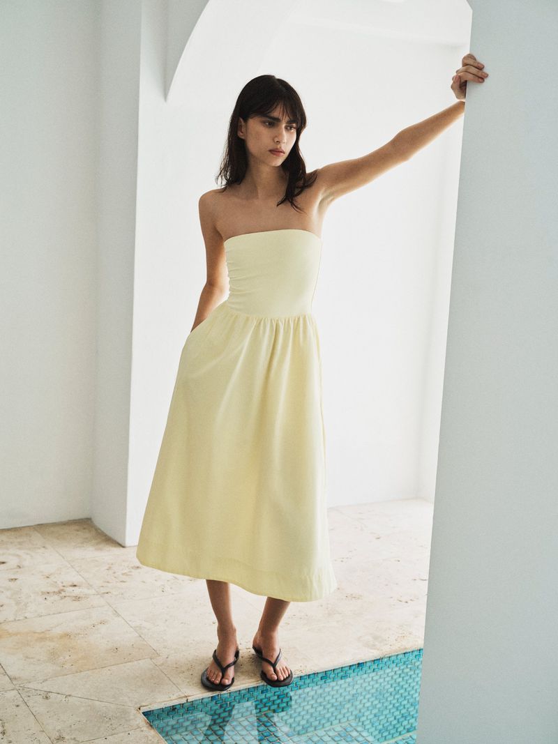 Woman wearing a strapless pale yellow midi dress and black sandals, leaning against a white doorway beside a tiled water feature.