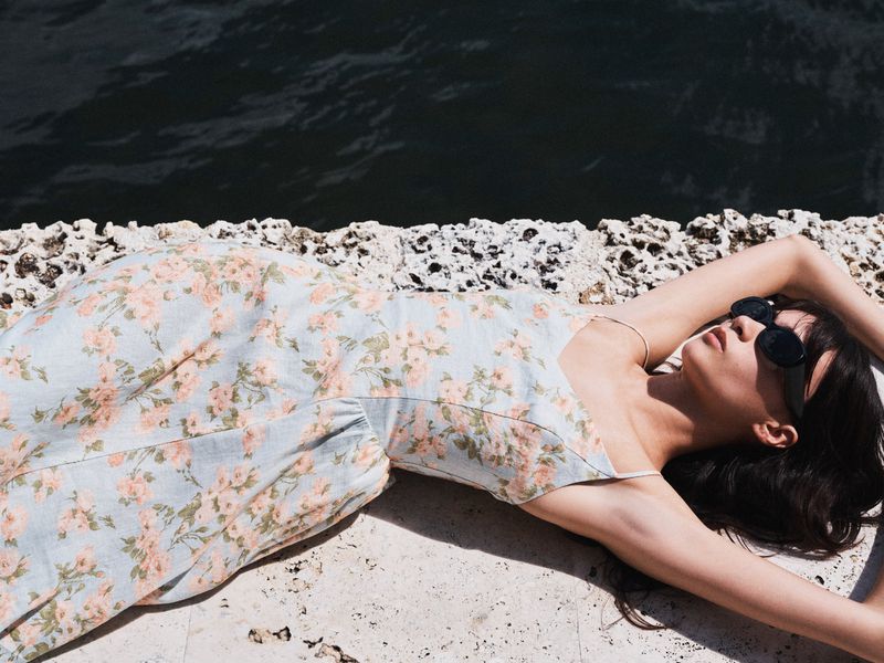 Woman wearing a floral slip dress and black sunglasses lying on a textured stone ledge beside dark open water.