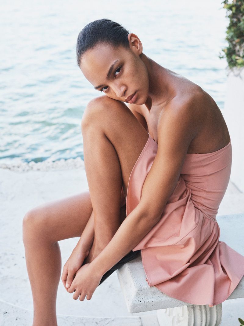 Model wearing a strapless blush pink dress seated on a stone bench overlooking the ocean in natural light.