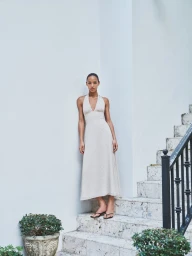 A woman in an ivory halter midi dress stands on stone outdoor steps beside a white wall and potted plant.