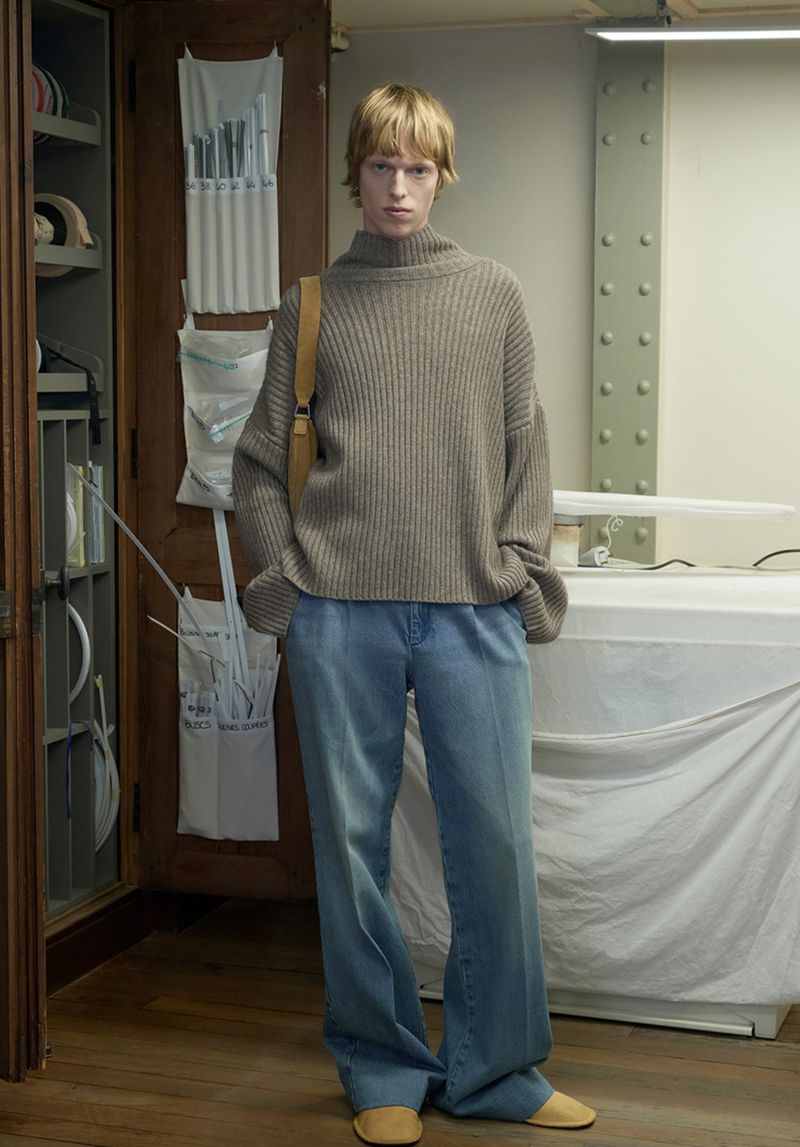 Model Joseph Uyttenhove wearing a taupe ribbed turtleneck sweater and wide-leg denim jeans standing in an atelier studio space.