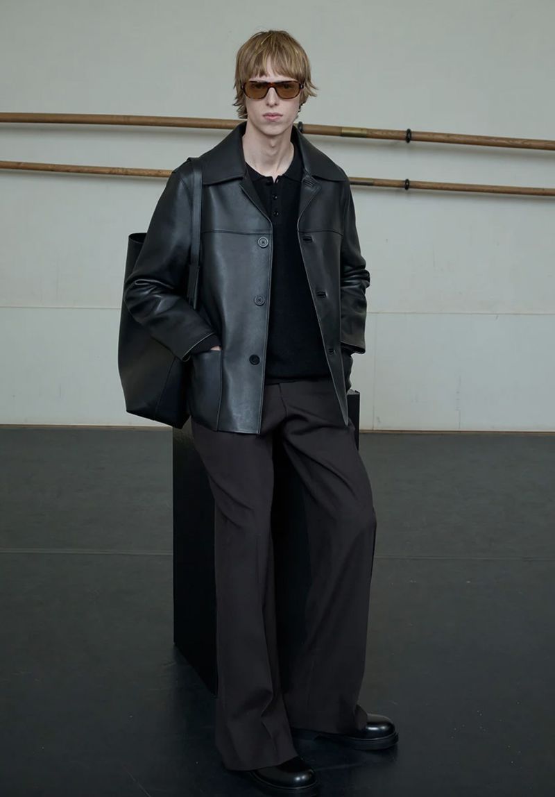 Male model in black leather jacket and dark trousers standing against an interior wall with wooden ballet barres