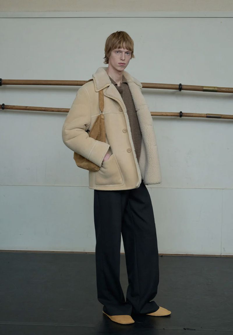 Model Joseph Uyttenhove wearing a beige shearling coat, brown sweater, and black trousers next to wooden ballet barres.