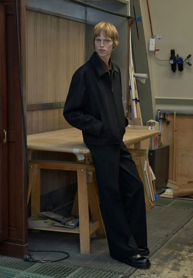 Model Joseph Uyttenhove in a black zip jacket and matching wide-leg trousers leaning against a wooden table for Sandro.