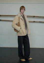 Model Joseph Uyttenhove wearing a beige shearling coat, brown sweater, and black trousers next to wooden ballet barres.