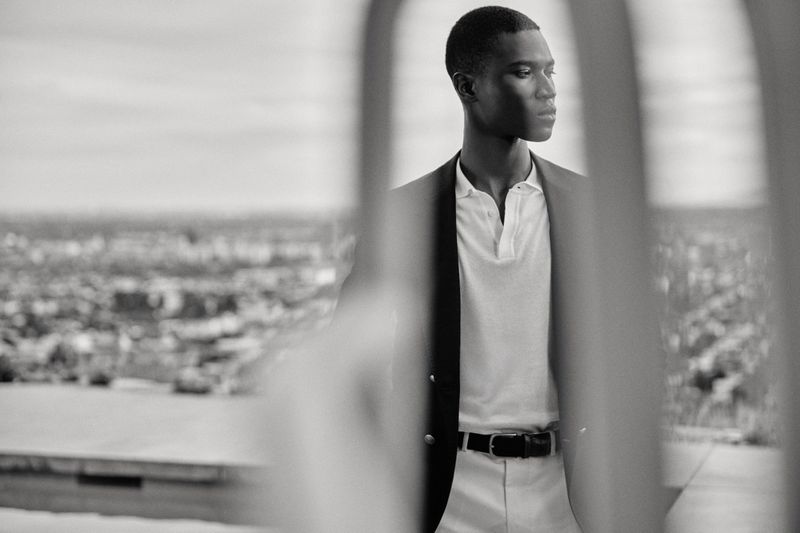Black and white portrait of a Black male model wearing a dark blazer and white polo, seen through blurred foreground bars.
