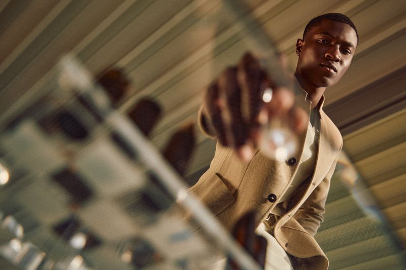Kwaku Ansong wearing a camel tailored jacket reaching toward a glass chessboard from a low angle for Paul Stuart.