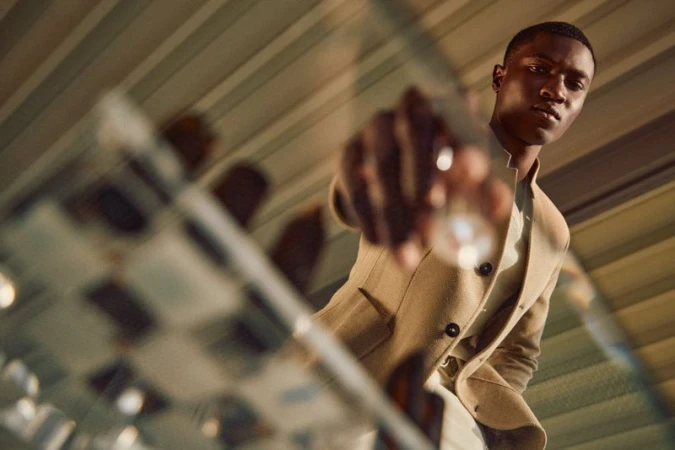 Kwaku Ansong wearing a camel tailored jacket reaching toward a glass chessboard from a low angle for Paul Stuart.