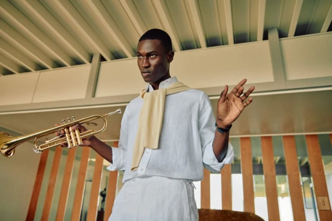 Model Kwaku Ansong holding a brass trumpet in a white linen shirt and trousers with a sweater draped over his shoulders.