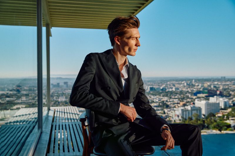 Model Finnlay Davis in a dark double-breasted suit seated on a modernist balcony overlooking an expansive cityscape.