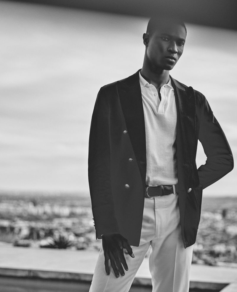 Monochrome portrait of model Kwaku Ansong wearing a double-breasted blazer, polo shirt, and light trousers outdoors.