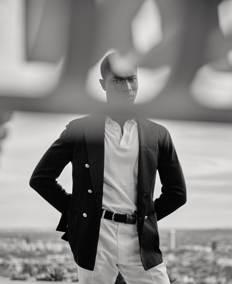 Black and white shot of an adult man wearing a jacket and pants, standing outdoors against a blurred urban background.