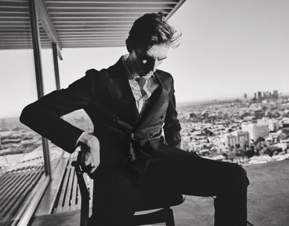 Model Finnlay Davis wearing a dark double-breasted suit seated before an expansive city skyline in a black and white portrait.
