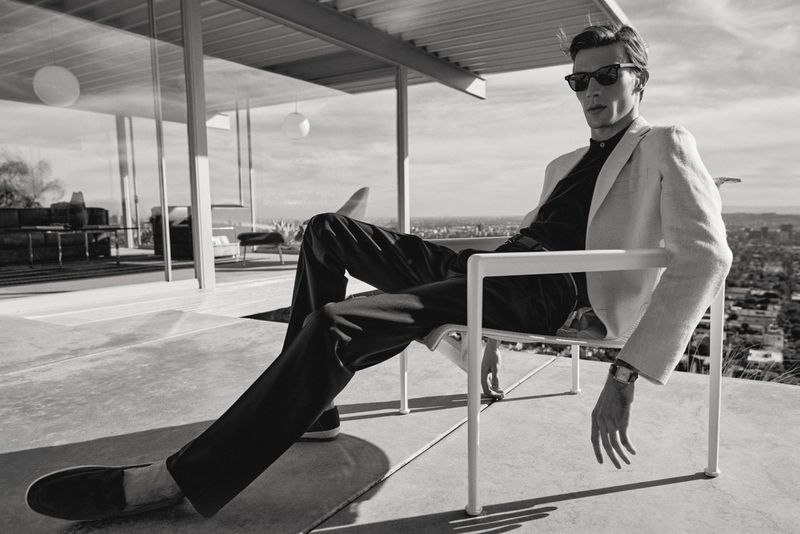 Monochrome fashion lookbook image of a male model wearing sunglasses and a light jacket seated on a modern terrace chair against a city view.