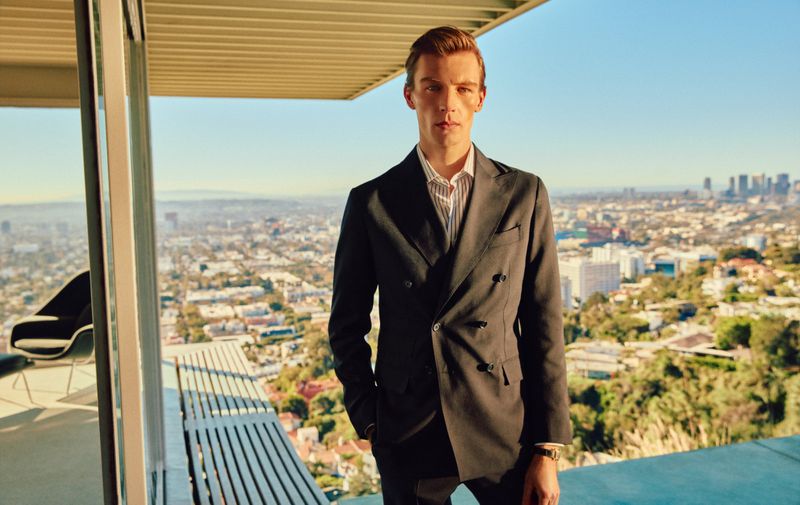 Model Finnlay Davis wearing a dark double-breasted suit jacket on a modernist terrace overlooking a sprawling city skyline.