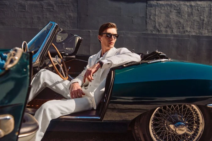 Finnlay Davis wearing a white tailored suit and sunglasses seated inside a classic green convertible sports car.