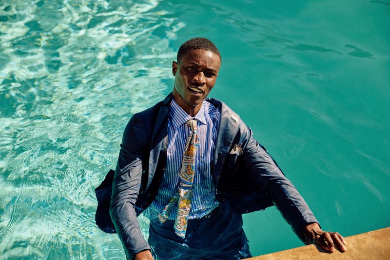 A Black man in a blue tailored suit and patterned tie wading in a turquoise outdoor swimming pool.
