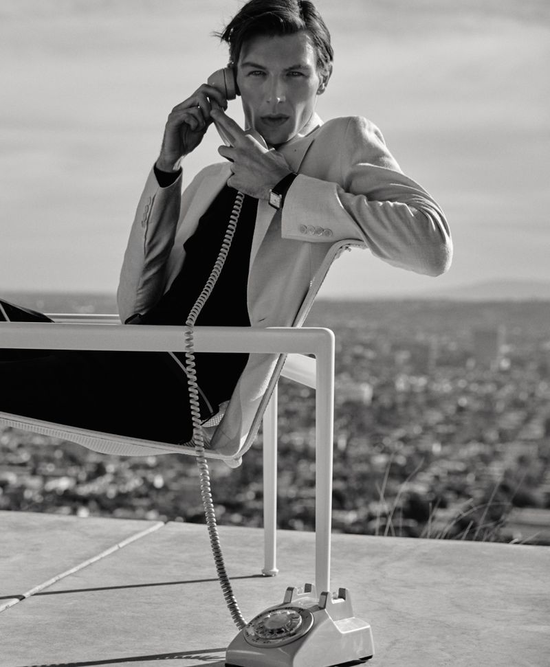 Black and white photo of Finnlay Davis in a light suit jacket seated outdoors holding a vintage rotary phone receiver to his ear.