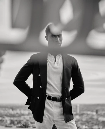 Black and white portrait of a Black man wearing a double-breasted blazer and polo shirt, framed through an out-of-focus foreground.