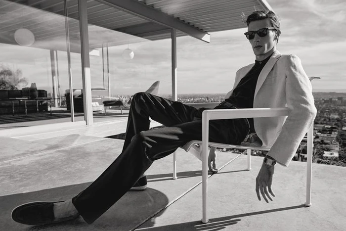Monochrome fashion lookbook image of a male model wearing sunglasses and a light jacket seated on a modern terrace chair against a city view.