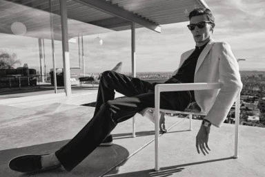 Monochrome fashion lookbook image of a male model wearing sunglasses and a light jacket seated on a modern terrace chair against a city view.