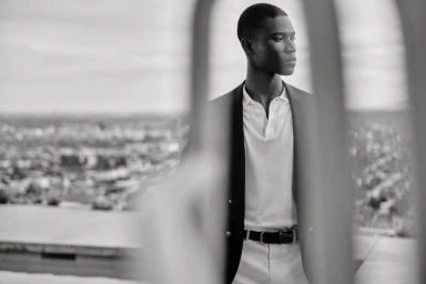 Black and white portrait of a Black male model wearing a dark blazer and white polo, seen through blurred foreground bars.