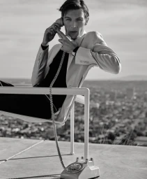 Black and white photo of Finnlay Davis in a light suit jacket seated outdoors holding a vintage rotary phone receiver to his ear.