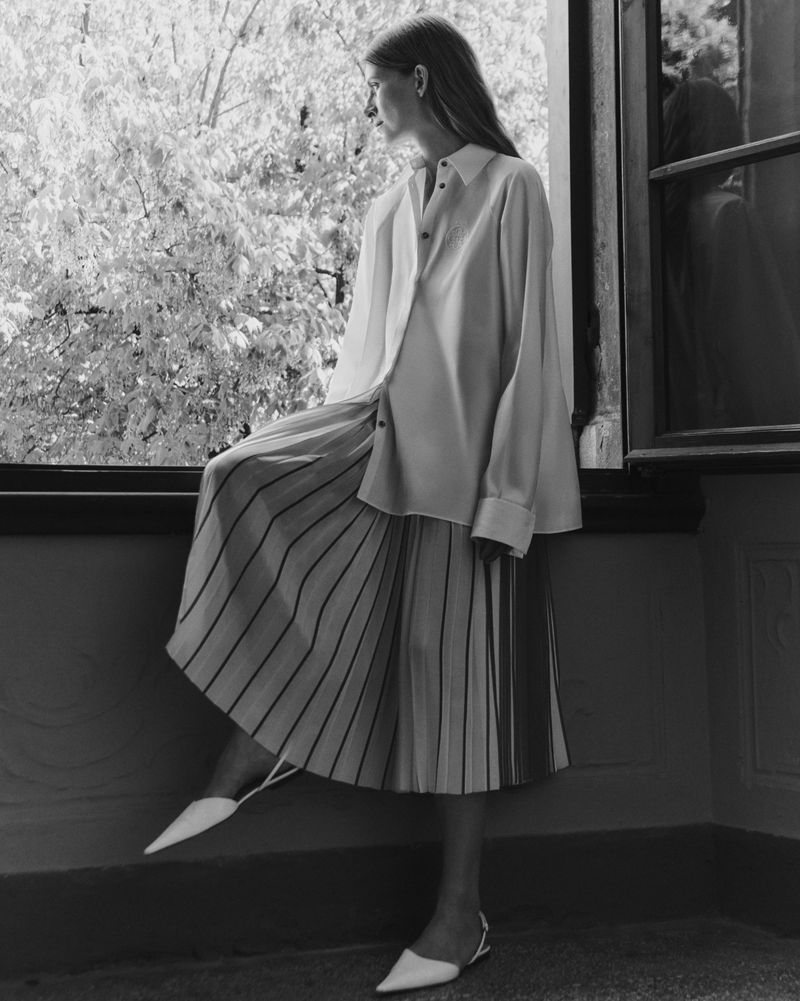 Model Luca Gajdus wearing a pleated skirt and white shirt by Giada standing by a window in a black and white photograph