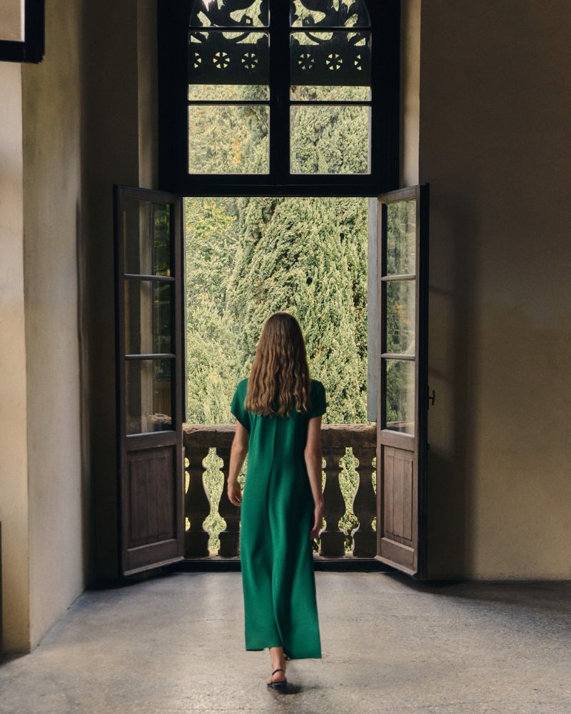 Woman in a flowing green maxi dress seen from behind walking toward open balcony doors with a balustrade overlooking lush green trees.