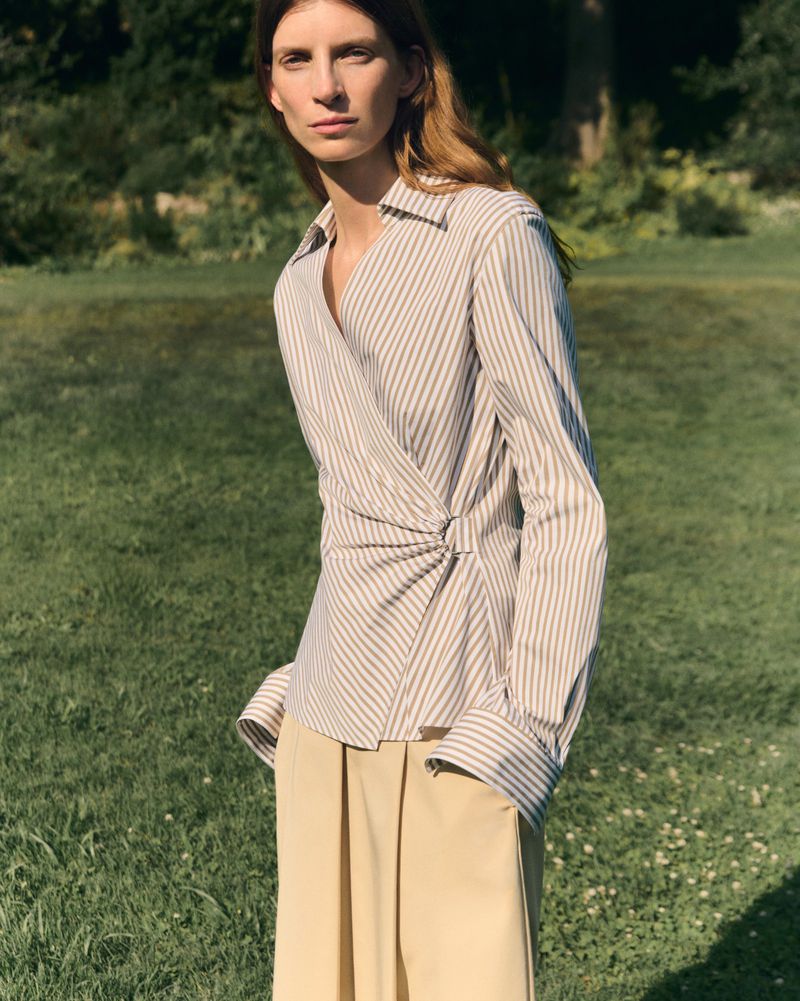 Model Luca Gajdus wearing a striped long-sleeve wrap blouse and beige pleated trousers on a grassy lawn outdoors.