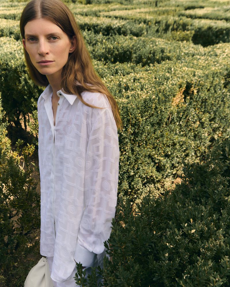 Model Luca Gajdus wears a sheer white patterned button-up shirt standing among green garden hedges for Giada.
