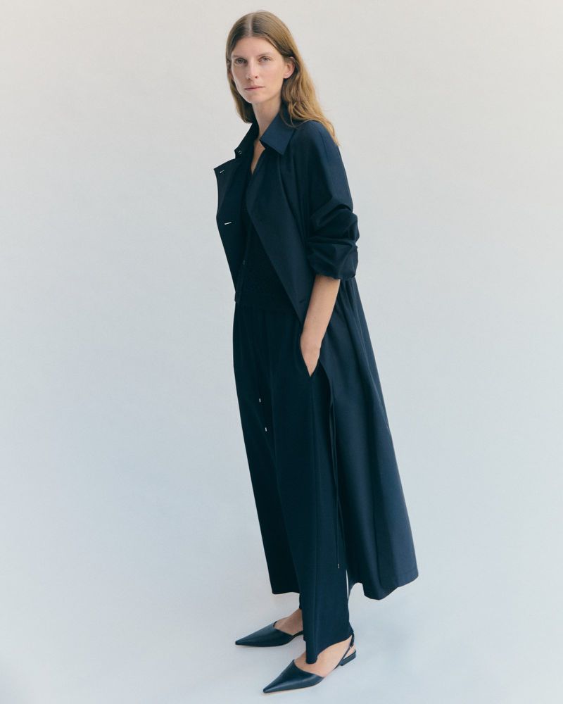 Model Luca Gajdus wearing a long dark navy duster coat and matching trousers with pointed flats against a white studio backdrop.