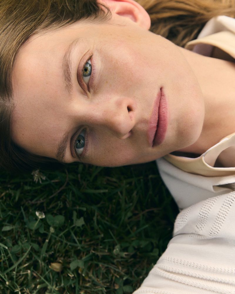 Close-up portrait of model Luca Gajdus lying on green grass wearing a white textured collared top.