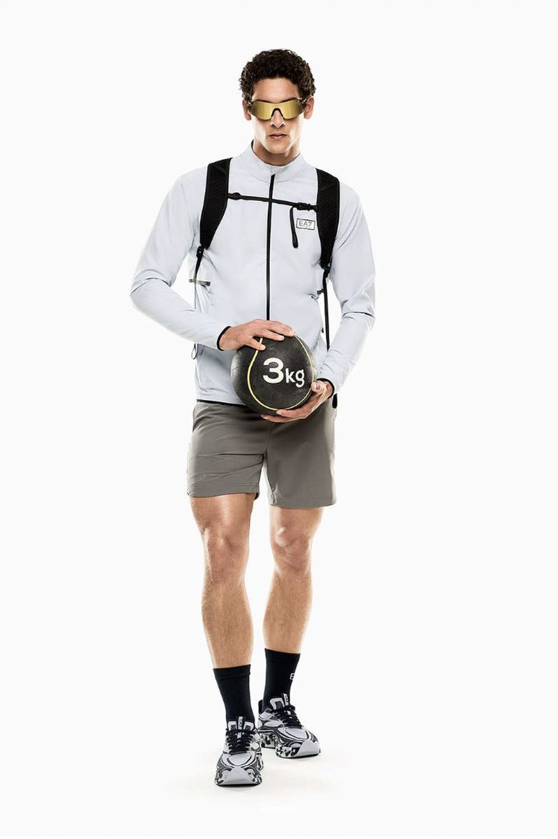 Athletic male model in a light grey zip jacket and shorts holding a black 3kg medicine ball against a white studio background.