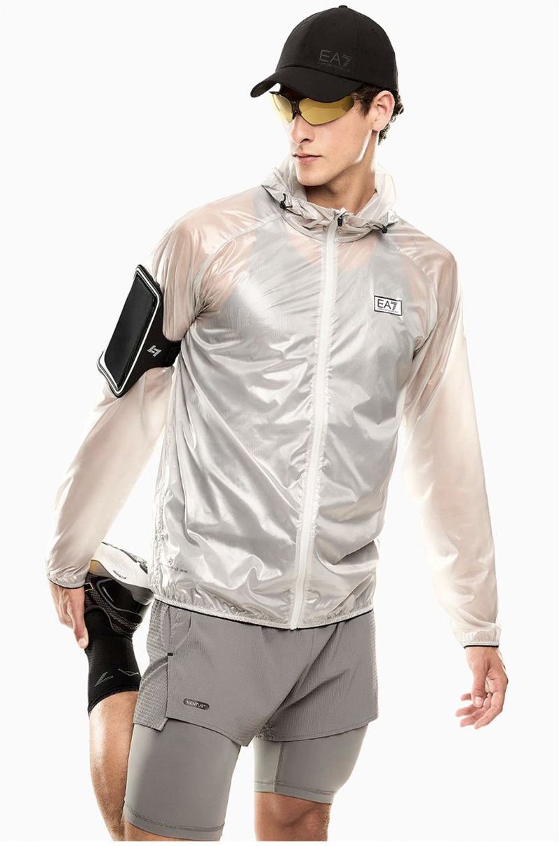 Male model stretching in a translucent running jacket, grey athletic shorts, black cap, and yellow sunglasses against a white background.