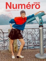 Model Daria Zolotova in a red and blue colorblock pullover and black shorts leans against a blue scenic telescope by a lake railing.