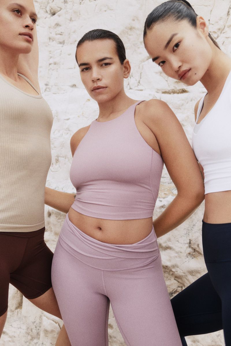 Three women in athletic wear posing against a light-colored background. The center model wears lilac leggings and a top, with other models flanking.