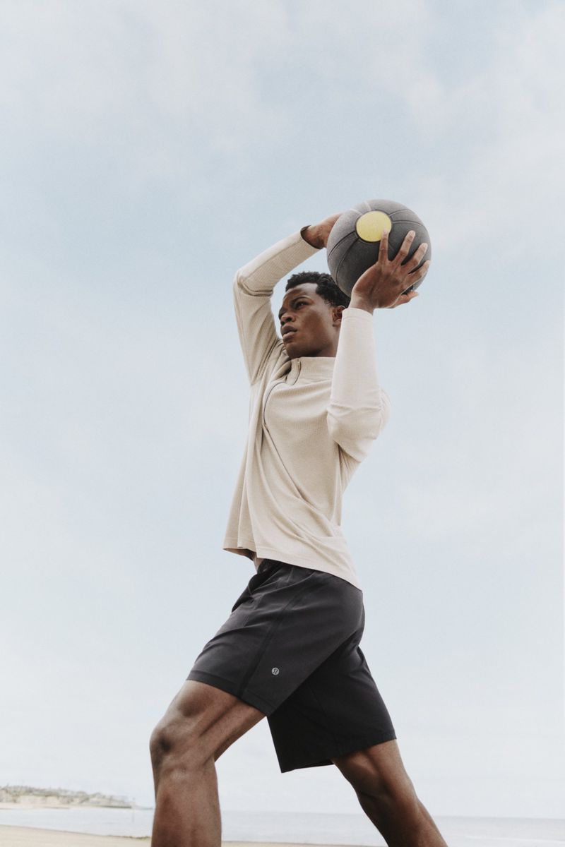 Athletic man outdoors in sportswear holds a medicine ball overhead, ready to throw, against a bright sky backdrop.
