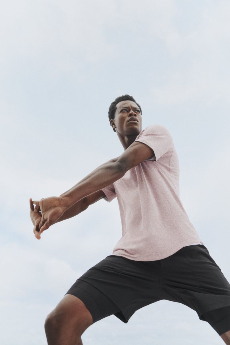 Athletic man stretching outdoors against a bright, overcast sky. He wears a t-shirt and athletic shorts. Low angle, upward view.