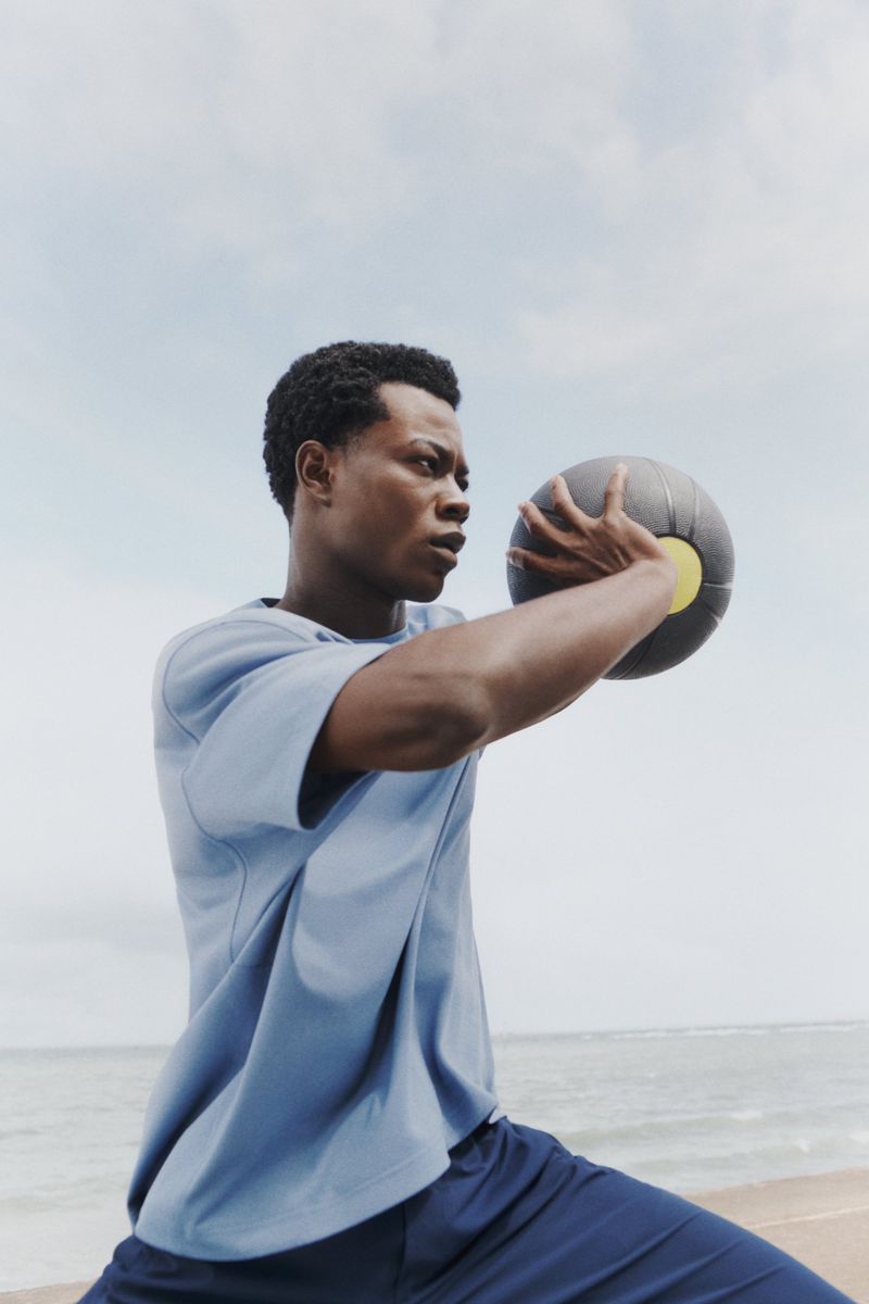 Athletic Black man works out on the beach with medicine ball. Wearing casual t-shirt and shorts against a background of the ocean and sky.