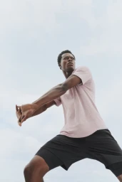 Low-angle shot of a Black man stretching his arms outdoors wearing a pale pink t-shirt and black athletic shorts against an open sky.