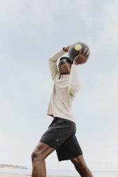 Man wearing a cream long-sleeve top and black shorts holding a medicine ball outdoors under an overcast sky