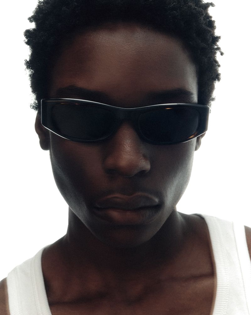 Close-up portrait of a Black model wearing dark rectangular sunglasses and a white ribbed tank top against a bright white background.