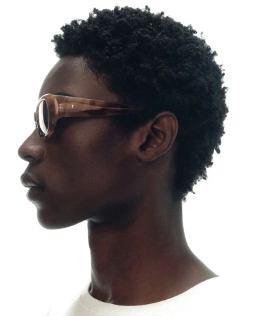Side profile of a Black model with short textured afro hair wearing brown tortoiseshell sunglasses against a bright white background.